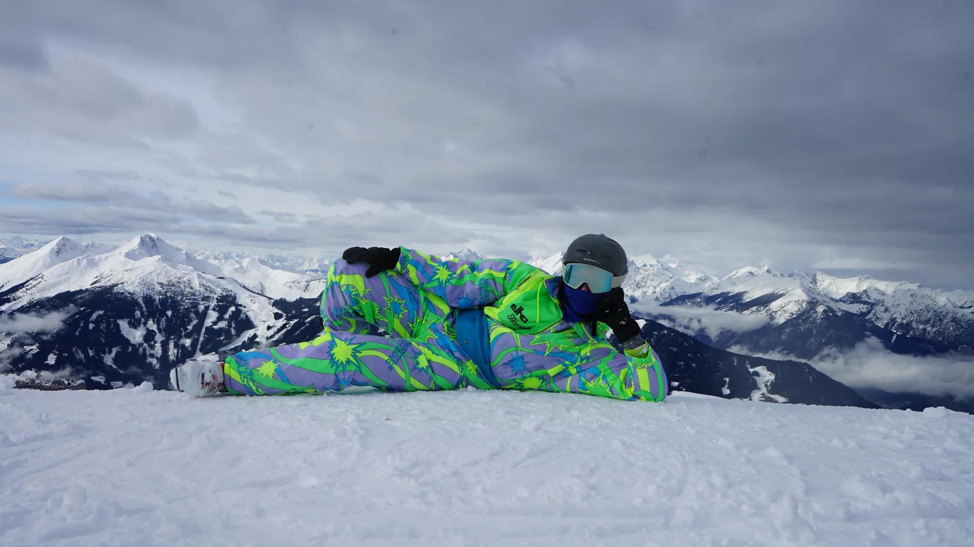 Person in farbigem Schneeanzug posiert vor einem winterlichen Bergpanorama